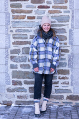 Full-length shot of cute smiling young woman in checkered winter coat leaning against stone wall in the old town Petit-Champlain sector, Quebec City, Quebec, Canada