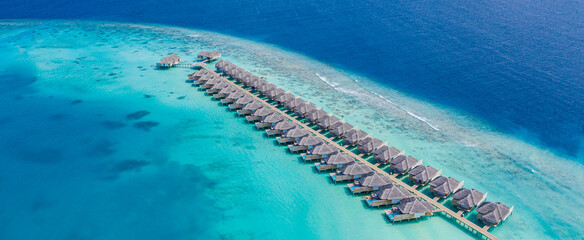Fototapeta premium Aerial view of Maldives island, luxury water villas resort and wooden pier. Beautiful sky and ocean lagoon beach background. Summer vacation holiday and travel concept. Paradise aerial landscape pano