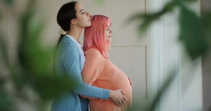 Woman touching her pregnant girlfriend belly and looking out through window