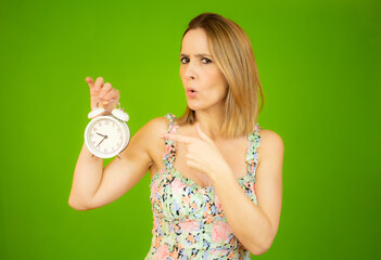 Young woman holding alarm clock standing over isolated green background pointing with hand and finger