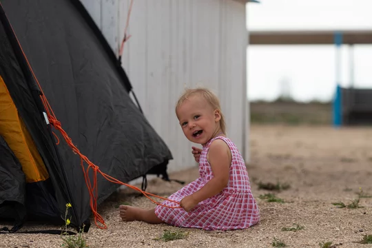 a little girl in a pink dress sits on the sand on the beach, next to a  tourist green tent, holding an orange rope from the tent in her hand and  smiling
