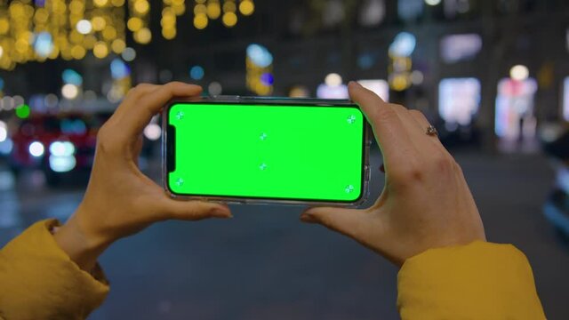 Woman hold smartphone with green screen. Background of busy bustling night city life, traffic, light and cars. Smartphone perfect for product placement and integration. Woman use phone for application