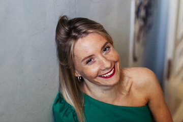 portrait of a beautiful young smiling woman with freckles in a green dress and with red lipstick standing leaning against a white wall