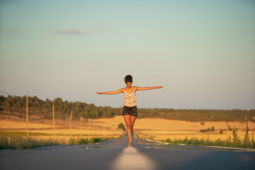 Fototapeta premium Young girl, walking in the middle of a lonely straight road, late in the day. With the hands stretched out in the shape of a cross, to maintain balance and not fall from the dividing line.