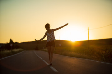 Young girl, walking in the middle of a lonely straight road, late in the day. With the hands stretched out in the shape of a cross, to maintain balance and not fall from the dividing line.