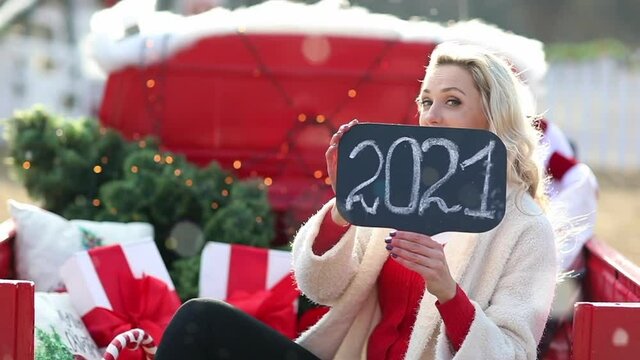 Woman posing in a car with Christmas tree and name plate with 2021 sign. Snow.