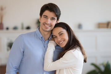 Obraz na płótnie Canvas Head shot portrait smiling man and woman hugging, standing at home, happy young couple posing for family photo together, positive wife and husband looking at camera, cuddling, enjoying leisure time