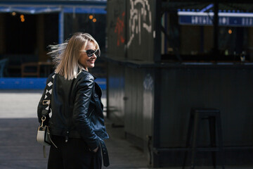 Obraz na płótnie Canvas young beautiful smiling stylish blonde woman in sunglasses, black pants and leather jacket posing on the don of buildings in an urban setting
