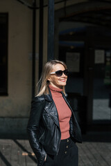Obraz premium young beautiful smiling stylish blonde woman in sunglasses, black pants and leather jacket posing on the don of buildings in an urban setting on sunny day