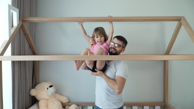 Young father with little daughter do gymnastics on horizontal bar of crib in form of house at home. Sport and gymnastics concept. Self-isolation. Father and child girl play active games.