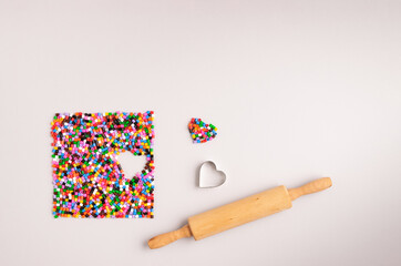 A rolling pin and a heart shaped cookie mold next to colorful plastic beads neatly arranged on a pastel grey background. Creative flat lay concept.