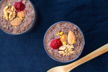Two glasses of chocolate, orange and chia seed pudding with nut and raspberry topping. Top view, dark blue background.