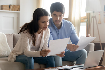 Close up serious young couple reading documents, calculating finances, sitting on couch at desk...