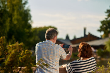 Back view mature man taking a photo on his smartphone. Adult senior couple enjoying the nature sitting outdoors on the grass.