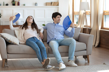 Exhausted man and woman waving blue paper fans, breathing, resting sitting on couch in living room,...
