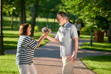 Fototapeta premium Happy woman recieving flowers from her husband. Mature mans giving gift to her mature wife. Green sunny park background.