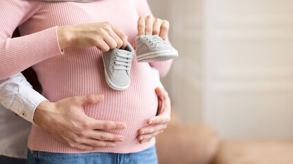 Unrecognizable Husband Holding Tiny Shoes Near Pregnant Wife's Belly Indoors