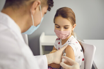 Brave girl in face mask looking at needle in doctor's hands while getting a flu shot at clinic or...