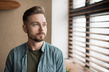 Serious thinking man looking at window in a house. Close up male portrait at home. Anxiety, stress, lockdown, mental health concept