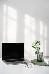 Workplace, home desk with laptop computer, glasses and notebook lying on white desk with a beautiful spring flower at background.