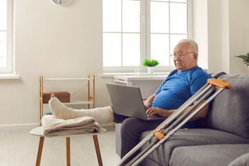 Sad senior man with broken leg in cast sitting on sofa at home and using laptop computer....