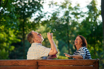 Retired couple blowing soap bubbles outdoors. Back view man and woman having fun on the park bench.