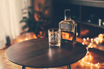 A glass and a bottle of whiskey scotch on a wooden table at home with cosy candid lights