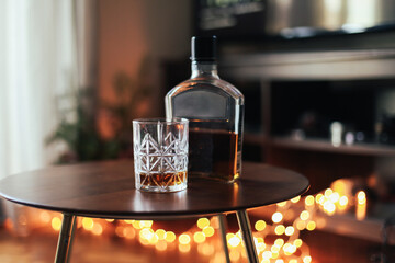 A glass and a bottle of whiskey on a wooden table at a cosy corner with candid lights