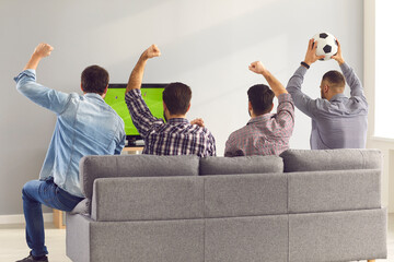 Four excited men friends sitting backwards on sofa at home and watching emotionally soccer football...