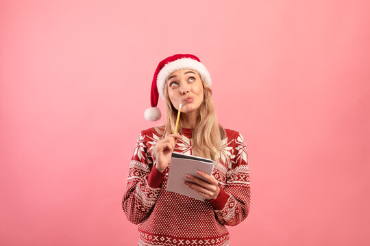 Charming young lady in Christmas outfit thinking about her New Year resolutions, writing wish list over pink background