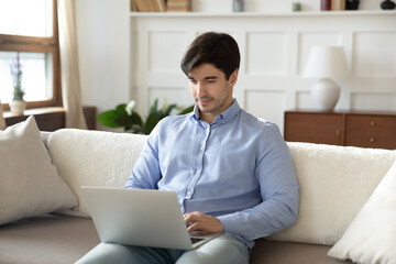 Focused man using laptop, sitting on cozy couch at home, looking at computer screen, chatting or shopping in internet, reading message, freelancer working online, spending leisure time with device