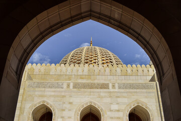 A white dome in contrast with the blue sky in Sultan Qaboos Grand Mosque. The scene is seen from behind an arch. Muscat, Oman.