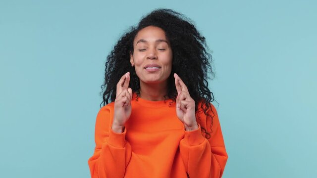 Pleading young african american woman 20s years old in orange sweatshirt hold hands folded in prayer begging about something making wish keep fingers crossed isolated on pastel blue color background