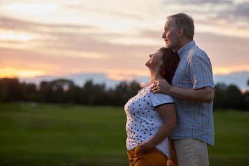 Mature man higs his wife from behind. Side view happy senior couple outdoors. Green field background.