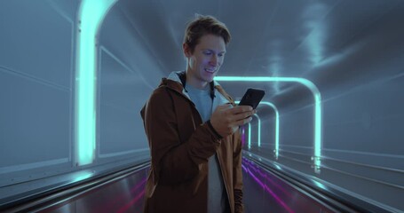 Cinematic portrait of young man travel through futuristic tunnel and check news and apps on his smartphone. Conceptual candid millennial man, commute in metro or airport.Technology information concept - Powered by Adobe