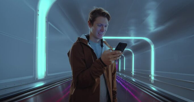 Cinematic portrait of young man travel through futuristic tunnel and check news and apps on his smartphone. Conceptual candid millennial man, commute in metro or airport.Technology information concept