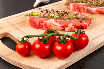 fresh chopped raw pork steaks with seasoning, ripe tomatoes and thyme on a cutting kitchen board on a black wooden table