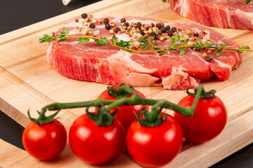 fresh chopped raw pork steaks with seasoning, ripe tomatoes and thyme on a cutting kitchen board on a black wooden table