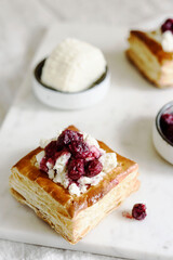 Puff pastry squares with ricotta and cherry sauce on white marble tray. Baked small square cakes topped with cheese and berry sauce