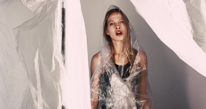 Female abuse. Woman protection. Domestic violence. Isolation safety. Tired assaulted lady wearing transparent polyethylene film suffering fainting on gray illuminated art background.