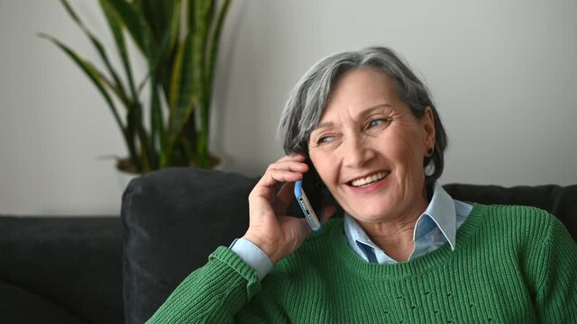 Portrait of charming grandmother wearing green sweater sitting on the sofa, friendly senior mature gray-haired lady is talking, listening to a caller, staying in touch with her family in pandemic