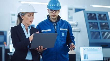 Female Manager and Project Engineer Wearing Hardhats Use Laptop in Industrial Factory, Talk, Plan Productivity Optimization. Production Line Workers Operate CNC Machinery, Program robot arm
