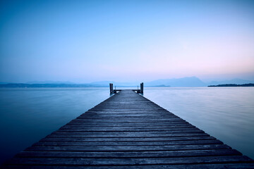 An old wooden pier extends into the clear blue waters of the lake. A solitary path towards the calm and peaceful silence of nature, with the sweet sound of the waves.