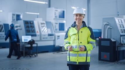Beautiful Smiling Female Engineer Wearing Safety Vest and Hardhat Holds Safety Goggles. Professional Woman Working in the Modern Manufacturing Factory. Facility with CNC Machinery and robot arm