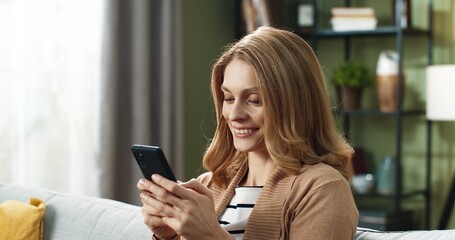 Close up of beautiful blonde woman sitting on sofa with smartphone in hands and laughing reading news online.