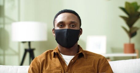 Fototapeta na wymiar Close up portrait of young african american man sitting at home on sofa in protective mask and looking at camera smiling.