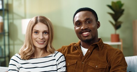 Portrait of happy multinational couple sitting at home on couch hugging looking at each other,...