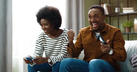 Young happy african american couple sitting at home on couch and playing video games on console. Happy man dances sitting, rejoicing in victory.