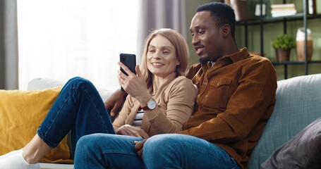 Beautiful multinational couple lying at home on couch and discussing news from social networks. Attractive woman holding phone in hands.