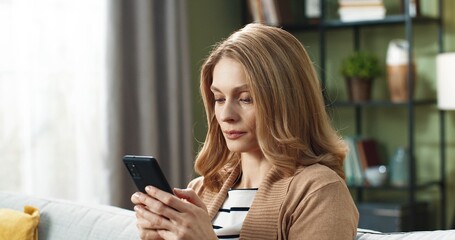 Close up of beautiful blonde woman sitting at home on couch and typing message on smartphone looks up and smiles at camera.